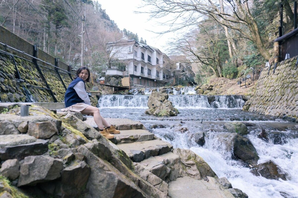 An Asian woman relaxes on stone steps by a river with rapids, village in background.